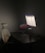 A dimly lit workspace with a dark wooden desk, featuring a modern ergonomic chair with a mesh backrest and lumbar support cushion. A large monitor displays a light-colored application interface, and a laptop with a colorful desktop background is positioned slightly open. On the desk are a keyboard, a mouse, a cylindrical container, and a small plant in a pot.