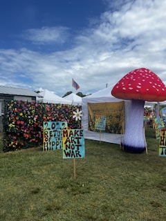 A vibrant outdoor setting with colorful tents and artistic displays. Tall, inflatable mushroom art piece is prominent on the right. Signs with phrases like '2022 Return of the Hippie' and 'Make Art Not War' are placed on the grass. A lush flower wall decorates the area. The sky is partly cloudy, adding a bright backdrop.