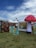 A vibrant outdoor setting with colorful tents and artistic displays. Tall, inflatable mushroom art piece is prominent on the right. Signs with phrases like '2022 Return of the Hippie' and 'Make Art Not War' are placed on the grass. A lush flower wall decorates the area. The sky is partly cloudy, adding a bright backdrop.