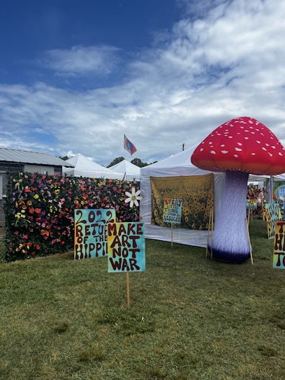A vibrant outdoor setting with colorful tents and artistic displays. Tall, inflatable mushroom art piece is prominent on the right. Signs with phrases like '2022 Return of the Hippie' and 'Make Art Not War' are placed on the grass. A lush flower wall decorates the area. The sky is partly cloudy, adding a bright backdrop.