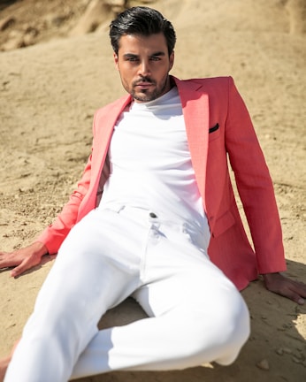 A man dressed in a light coral shirt and beige pants, with subtle sea-inspired jewelry, posing near underwater-themed decorations.