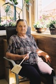 An elderly woman is seated in a chair near a window. She holds a fly swatter and is surrounded by potted plants. The sunlight streams in, casting shadows and highlighting her patterned shirt and dark pants.