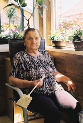 An elderly woman is seated in a chair near a window. She holds a fly swatter and is surrounded by potted plants. The sunlight streams in, casting shadows and highlighting her patterned shirt and dark pants.