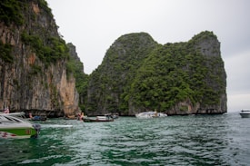 A scenic view of large, lush limestone cliffs covered in greenery rising from the water. Several boats, including long-tail and speedboats, are floating on the greenish-blue sea. The sky above is overcast, adding a muted tone to the landscape.