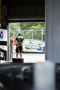 A person wearing a headset and a racing team shirt stands near a garage entrance, while a race car with logos and number decals speeds past on a track. The scene includes a bystander in an orange outfit, and green trees in the background.
