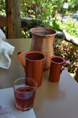 Several copper-colored mugs and a matching pitcher are arranged on a wooden table, with a glass of red liquid beside them. The background features a blurred garden scene filled with lush greenery and pink flowers, suggesting an outdoor setting.