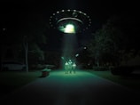 A dark outdoor setting illuminated by a large, glowing UFO hovering above. Three figures stand in its beam of light, surrounded by trees and parked cars. The scene is surreal and otherworldly, with an eerie green hue cast over the landscape.