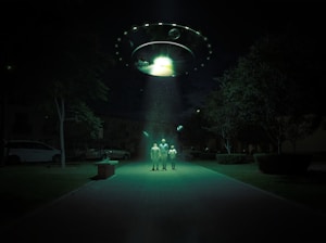 A dark outdoor setting illuminated by a large, glowing UFO hovering above. Three figures stand in its beam of light, surrounded by trees and parked cars. The scene is surreal and otherworldly, with an eerie green hue cast over the landscape.