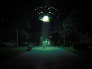 A dark outdoor setting illuminated by a large, glowing UFO hovering above. Three figures stand in its beam of light, surrounded by trees and parked cars. The scene is surreal and otherworldly, with an eerie green hue cast over the landscape.