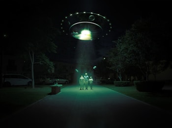 A dark outdoor setting illuminated by a large, glowing UFO hovering above. Three figures stand in its beam of light, surrounded by trees and parked cars. The scene is surreal and otherworldly, with an eerie green hue cast over the landscape.