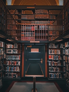 A richly wooden-paneled library alcove filled with shelves of colorful books. The scene is symmetrical, with a central green door labeled as Deck 46, indicating telephone books and city directories. The shelves surround the door, climbing up towards a mezzanine level, and are densely packed with books of various sizes and colors.