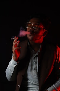 A person wearing glasses is exhaling smoke from a cigarette while sitting in a dark environment. They are dressed in a traditional or formal attire with a jacket over a light-colored shirt. The lighting is dim with a red hue highlighting one side of their face and hand.