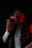 A person wearing glasses is exhaling smoke from a cigarette while sitting in a dark environment. They are dressed in a traditional or formal attire with a jacket over a light-colored shirt. The lighting is dim with a red hue highlighting one side of their face and hand.