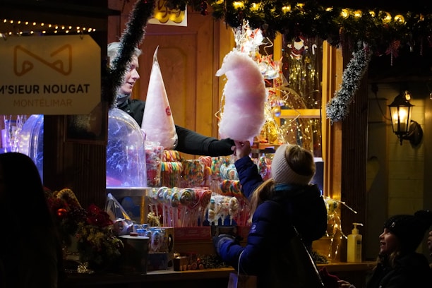 A festive market stall offers sweet treats, including colorful cotton candy and lollipops. A warm, wooden booth is adorned with holiday lights and decorations. People are engaged in purchasing sugary snacks, with a cheerful atmosphere prevailing.