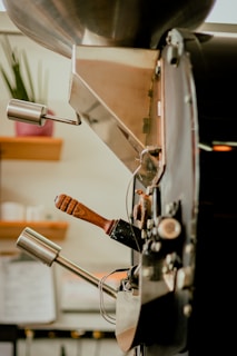 A close-up view of a coffee roasting machine with metallic levers and a wooden handle. The background features a blurred interior with a plant and shelves.
