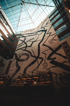 A tall, abstract mural with black geometric and organic shapes dominates a large wall in an indoor space. The ceiling is made of glass panels, allowing natural light to filter through. Below the mural, there are several pendant lamps with warm lighting, creating an inviting atmosphere.