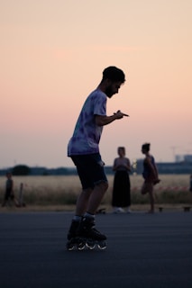 A person rollerblading in an open area with a soft pink and orange sunset in the background. The silhouette of the skater is captured mid-movement, wearing a tie-dye shirt and shorts. The scene includes other individuals in the background, with a field and some distant structures visible.