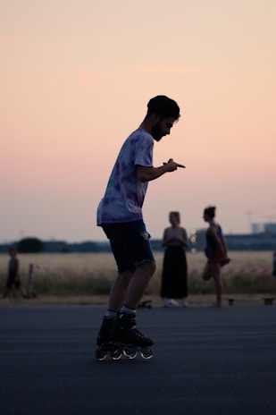 A person rollerblading in an open area with a soft pink and orange sunset in the background. The silhouette of the skater is captured mid-movement, wearing a tie-dye shirt and shorts. The scene includes other individuals in the background, with a field and some distant structures visible.