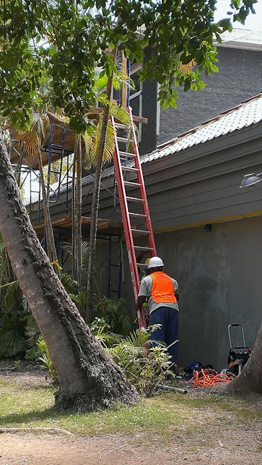 A worker wearing a safety vest and helmet stands near a ladder that is leaning against a building exterior. The scene is surrounded by palm trees and other greenery. There are construction tools and materials on the ground.