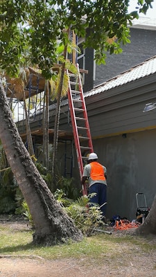 A worker wearing a safety vest and helmet stands near a ladder that is leaning against a building exterior. The scene is surrounded by palm trees and other greenery. There are construction tools and materials on the ground.
