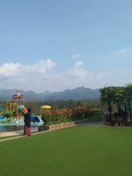 A playground with a colorful water slide sits in the foreground, surrounded by lush green grass and vibrant shrubbery. In the background, a mountain range creates a scenic backdrop under a clear blue sky with a few clouds. A person wearing a long dress and headscarf stands near the water slide.