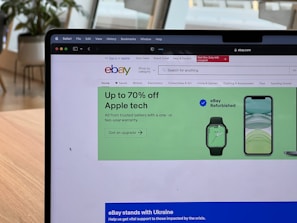 A laptop displays a web browser on the eBay homepage, showing a prominent green advertisement offering up to 70% off on Apple tech with a two-year warranty. The ad features images of a smartwatch and a smartphone. The surrounding environment includes a wooden table and blurred indoor plants in the background.