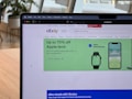 A laptop displays a web browser on the eBay homepage, showing a prominent green advertisement offering up to 70% off on Apple tech with a two-year warranty. The ad features images of a smartwatch and a smartphone. The surrounding environment includes a wooden table and blurred indoor plants in the background.
