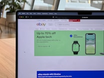 A laptop displays a web browser on the eBay homepage, showing a prominent green advertisement offering up to 70% off on Apple tech with a two-year warranty. The ad features images of a smartwatch and a smartphone. The surrounding environment includes a wooden table and blurred indoor plants in the background.