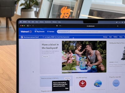 A laptop displaying the Walmart website is sitting on a wooden table. The website's interface shows a promotion for backyard activities, featuring a man and a child playing with water toys. There are promotional sections for rollbacks, savings, and July 4th preparations. The background includes a modern room with plants and stylish furniture.