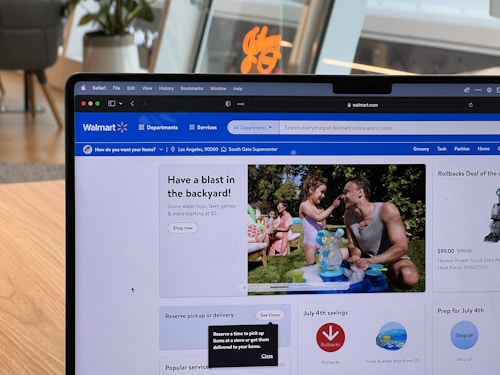 A laptop displaying the Walmart website is sitting on a wooden table. The website's interface shows a promotion for backyard activities, featuring a man and a child playing with water toys. There are promotional sections for rollbacks, savings, and July 4th preparations. The background includes a modern room with plants and stylish furniture.