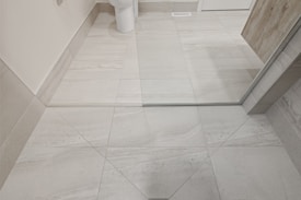 A bathroom setting featuring light gray tiled flooring. A partial view of a white toilet is visible near the corner. There is a glass shower door with metal trim and a section of a wooden plank wall. The tiles have a subtle pattern with light and dark lines.