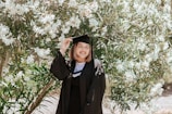 A person wearing a graduation cap and gown stands in front of a lush tree with white flowers, smiling warmly.