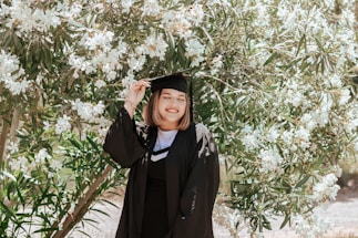 A person wearing a graduation cap and gown stands in front of a lush tree with white flowers, smiling warmly.