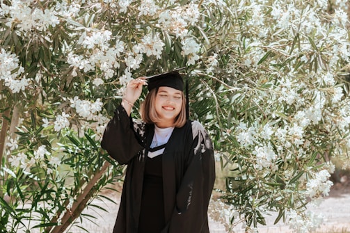 A person wearing a graduation cap and gown stands in front of a lush tree with white flowers, smiling warmly.