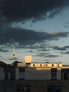 There is a building with modern architecture in the foreground, appearing under partly cloudy skies. A wind turbine is visible in the background, suggesting an emphasis on renewable energy.