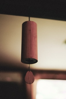 A wooden wind chime hangs indoors, with soft focus on the background creating a serene atmosphere. The cylindrical chime body is suspended from a thin string, with a teardrop-shaped piece below.