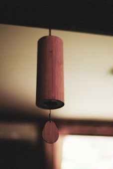 A wooden wind chime hangs indoors, with soft focus on the background creating a serene atmosphere. The cylindrical chime body is suspended from a thin string, with a teardrop-shaped piece below.