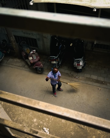 A person is sweeping a paved street. They are wearing a blue shirt and jeans, and there are a few scooters parked nearby. The perspective is from above, looking down through a metal railing.