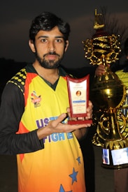 A man holds a red trophy with gold detailing and text on it, wearing a brightly colored sports jersey. Another larger gold trophy is partially visible in the background. The setting appears to be outdoors at dusk.
