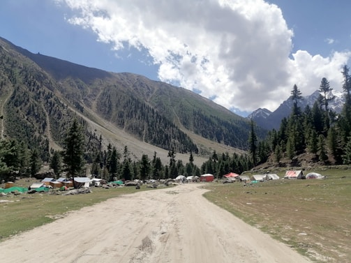 A dirt road traverses through a mountainous landscape with pine trees and slopes covered in green vegetation. Several tents of varying colors, including white, green, and red, are set up along the side of the road. The sky is partly cloudy with patches of blue visible.