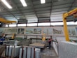 A large industrial workshop with high ceilings and metal beams. Various machines and equipment are present, likely used for cutting stone slabs. There are several large posters of architectural projects displayed on the wall. Two workers are visible, both wearing safety gear.