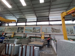 A large industrial workshop with high ceilings and metal beams. Various machines and equipment are present, likely used for cutting stone slabs. There are several large posters of architectural projects displayed on the wall. Two workers are visible, both wearing safety gear.