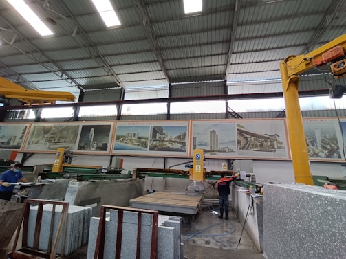 A large industrial workshop with high ceilings and metal beams. Various machines and equipment are present, likely used for cutting stone slabs. There are several large posters of architectural projects displayed on the wall. Two workers are visible, both wearing safety gear.