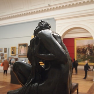 A dark, expressive sculpture of a human figure is displayed prominently in an art gallery. In the background, framed paintings are mounted on the walls, and several people are observing the artworks. The setting appears spacious with high ceilings and a large archway.