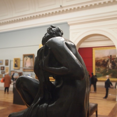 A dark, expressive sculpture of a human figure is displayed prominently in an art gallery. In the background, framed paintings are mounted on the walls, and several people are observing the artworks. The setting appears spacious with high ceilings and a large archway.
