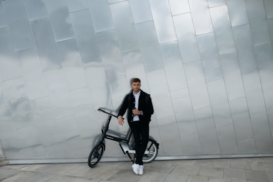 A person leans casually against a sleek, modern bicycle in an urban setting. The individual is dressed in a black jacket, white shirt, and white sneakers. The background features a reflective, metallic wall composed of rectangular panels, adding a contemporary feel to the scene.