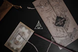 A collection of Harry Potter themed items, including a Marauder's Map, a Platform 9 3/4 train ticket, and a Deathly Hallows necklace. The map is detailed with a castle illustration and adorns black and brown tones. The train ticket is embellished with ornate designs and a vintage appearance. A wooden wand lies next to the items, partially covered by the chain of the necklace.