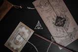 A collection of Harry Potter themed items, including a Marauder's Map, a Platform 9 3/4 train ticket, and a Deathly Hallows necklace. The map is detailed with a castle illustration and adorns black and brown tones. The train ticket is embellished with ornate designs and a vintage appearance. A wooden wand lies next to the items, partially covered by the chain of the necklace.