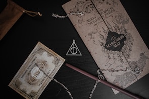 A collection of Harry Potter themed items, including a Marauder's Map, a Platform 9 3/4 train ticket, and a Deathly Hallows necklace. The map is detailed with a castle illustration and adorns black and brown tones. The train ticket is embellished with ornate designs and a vintage appearance. A wooden wand lies next to the items, partially covered by the chain of the necklace.