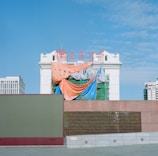A partially constructed or renovated building draped with colorful tarps, including orange, blue, and striped sheets. The building features traditional architectural elements and has red signage on top against a clear blue sky. Modern urban structures are visible in the background.
